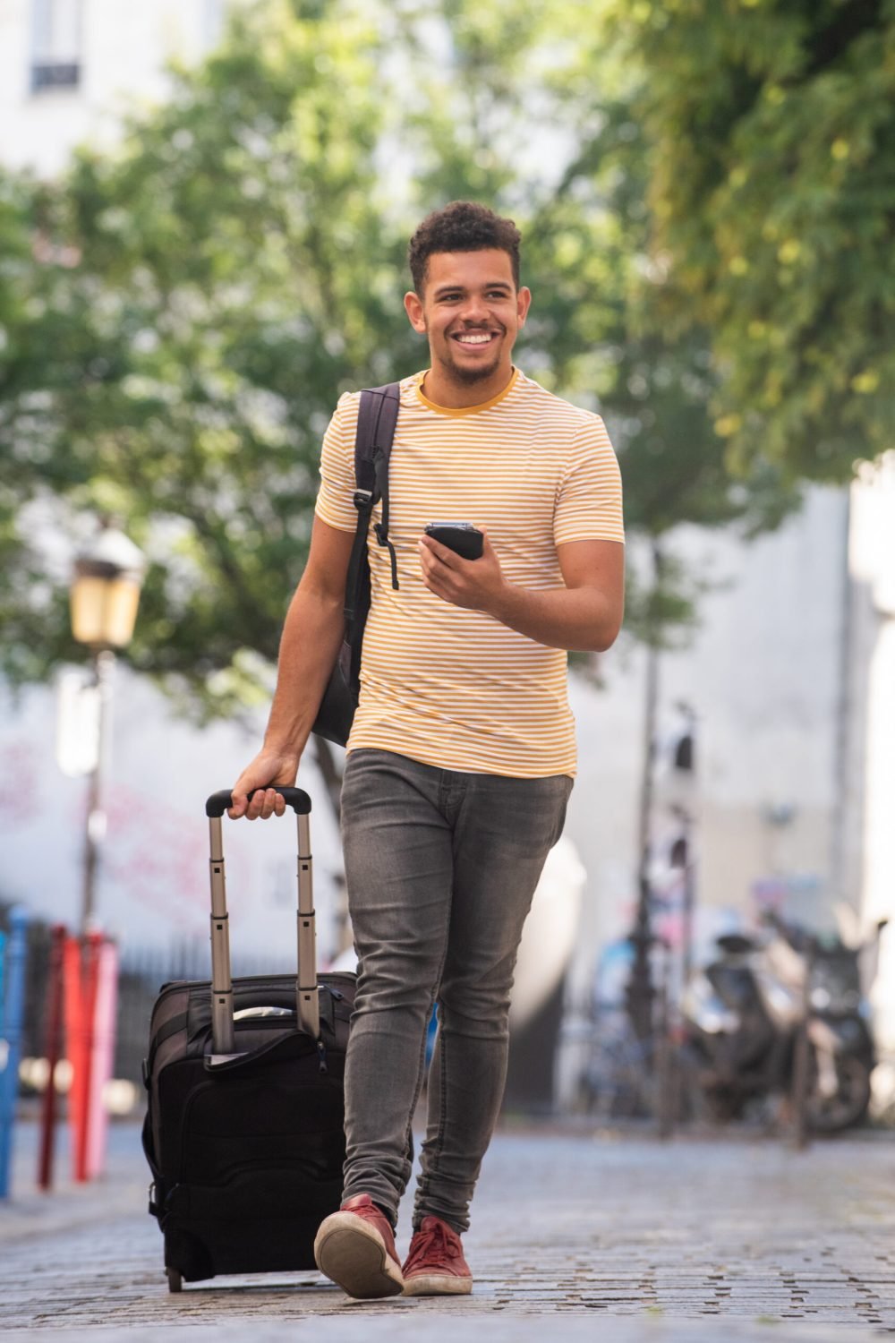 Full body portrait happy young man travel with cellphone and suitcase on street